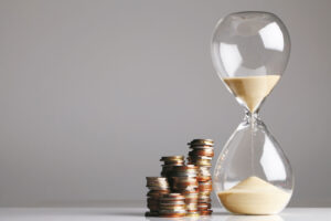 Das Bild zeigt eine Sanduhr, neben der mehrere kleine Münzstapel liegen. Der Sand rinnt langsam von der oberen in die untere Hälfte der Uhr und symbolisiert den Ablauf von Zeit. Die Darstellung vermittelt die Verbindung von Zeit und Geld – etwa im Sinne von Sparen, Vorsorge oder langfristigem Vermögensaufbau.