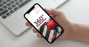 Smartphone in der Hand vor einem Laptop, auf dem Bildschirm ist das digitale Kundenmagazin ‚360° – Das Kundenmagazin der RVM Gruppe‘ zu sehen. Titelbild zeigt eine Person mit rotem Regenschirm auf einem Zebrastreifen. Modernes, professionelles Layout.