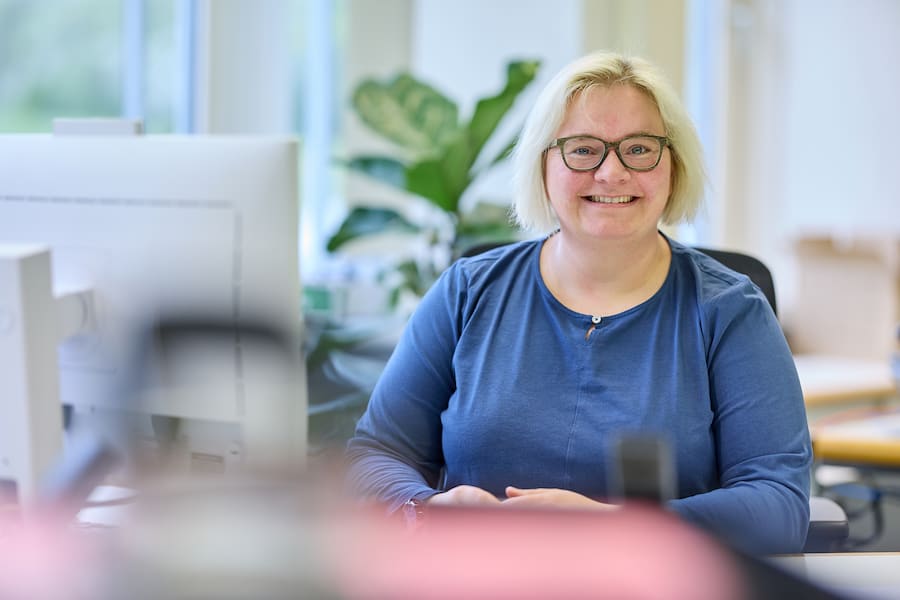 Lächelnde Frau mit blonden Haaren und Brille sitzt freundlich an ihrem Büroarbeitsplatz vor einem Computerbildschirm.