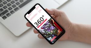 Smartphone in der Hand vor einem Laptop, auf dem Bildschirm ist das digitale Kundenmagazin ‚360° – Das Kundenmagazin der RVM Gruppe‘ zu sehen. Titelbild zeigt eine Person mit rotem Regenschirm auf einem Zebrastreifen. Modernes, professionelles Layout.
