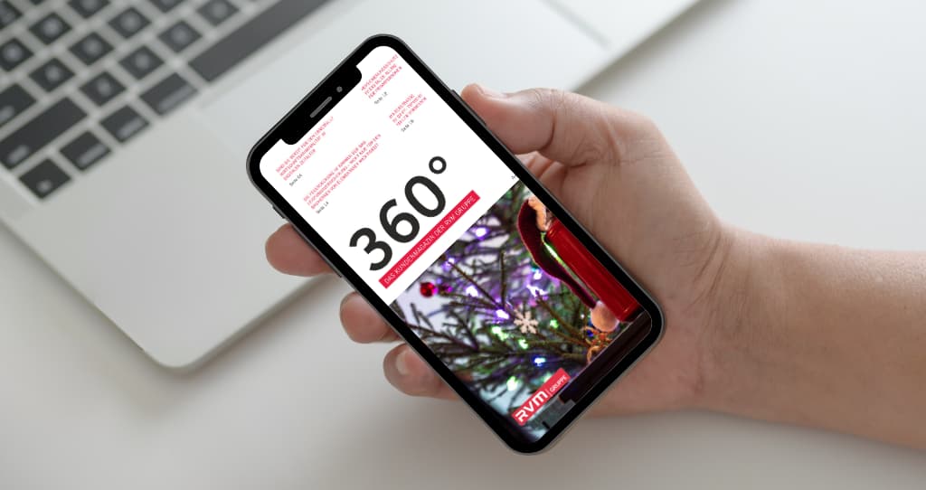 Smartphone in der Hand vor einem Laptop, auf dem Bildschirm ist das digitale Kundenmagazin ‚360° – Das Kundenmagazin der RVM Gruppe‘ zu sehen. Titelbild zeigt eine Person mit rotem Regenschirm auf einem Zebrastreifen. Modernes, professionelles Layout.